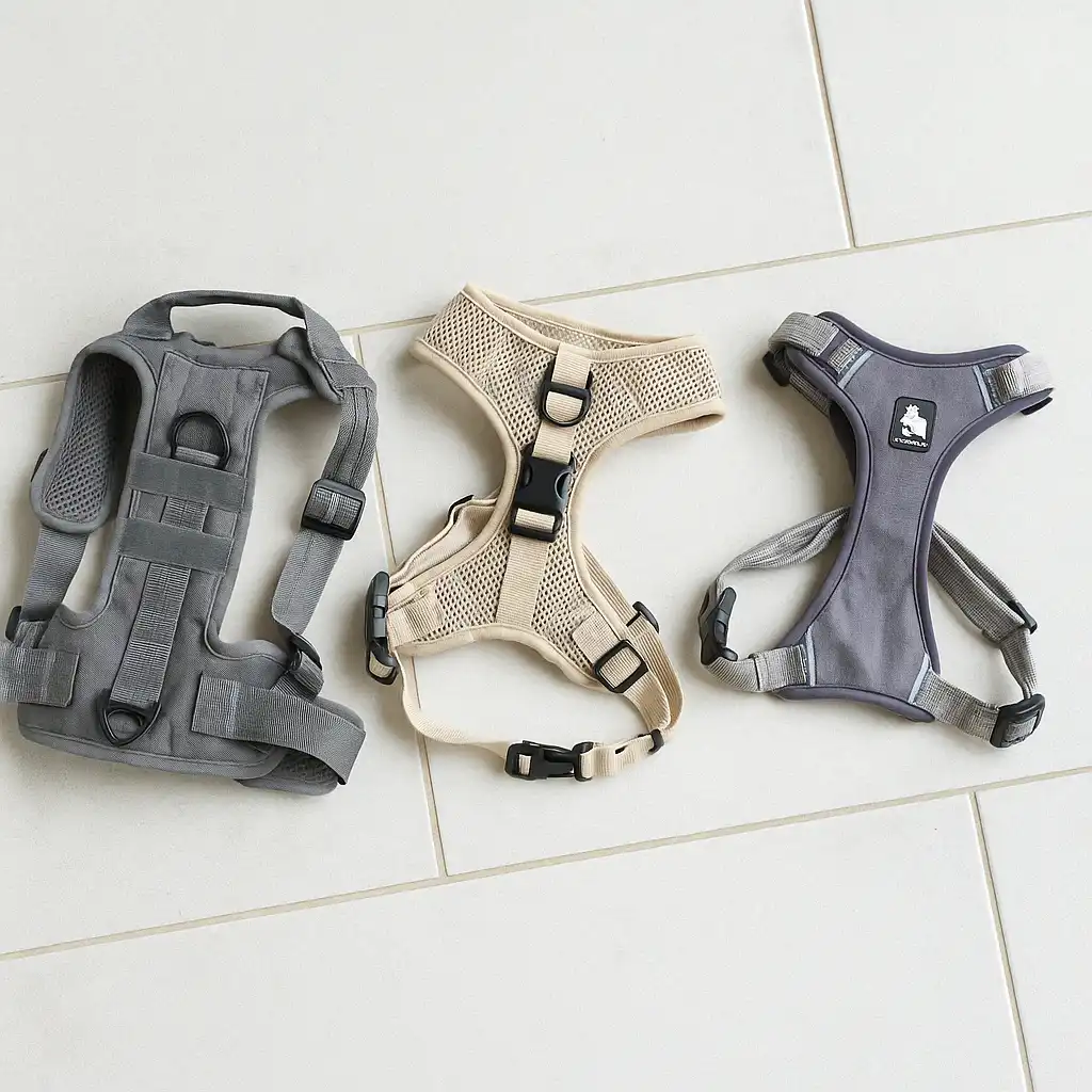 Three different dog harnesses laid out neatly on a wooden surface, showcasing a gray tactical vest, a sand-colored mesh harness, and a gray reflective front-clip harness, ideal for small dogs like pugs.